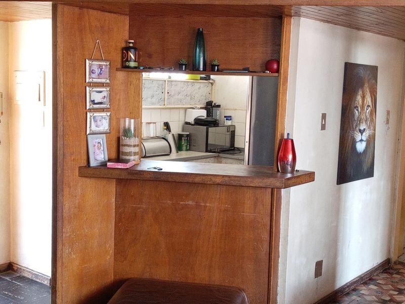 Open plan living area featuring a wooden serving hatch and breakfast bar connecting to the kitchen, with dated wood paneling and vinyl flooring.