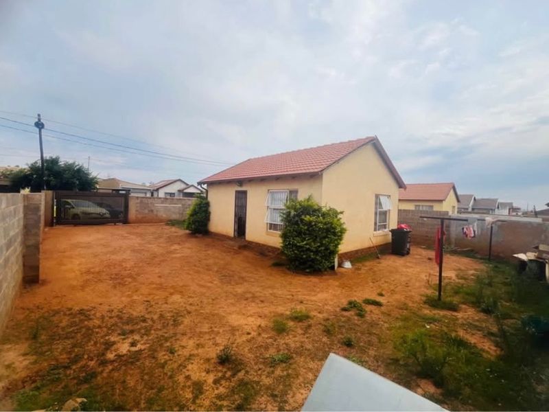 Rear view of a detached house featuring a tiled roof, security gate, and a large, unpaved yard enclosed by a perimeter wall.