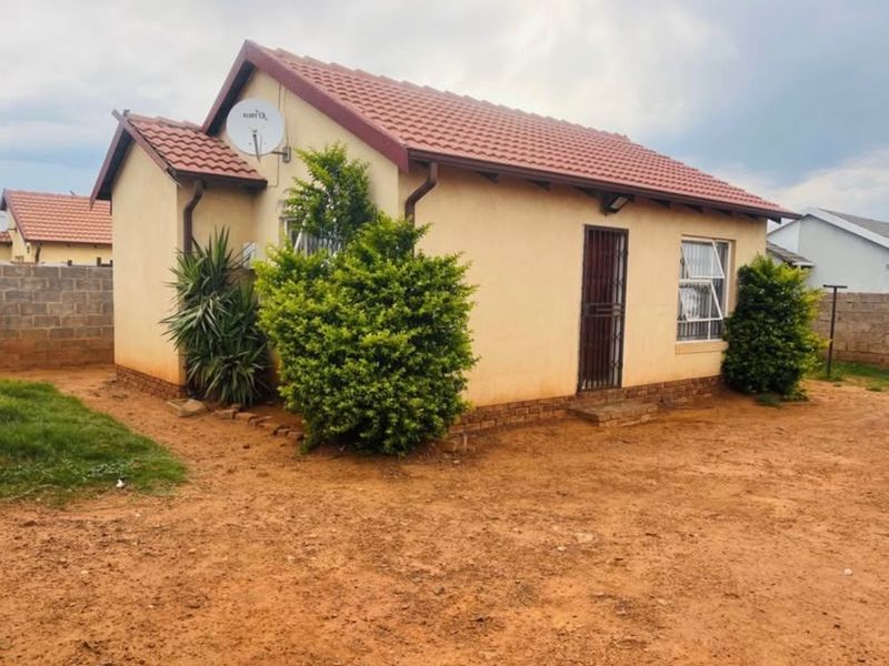 A detached single-story house featuring a tiled roof, security gate, and aluminium window frames, set on a spacious plot with an unpaved yard.