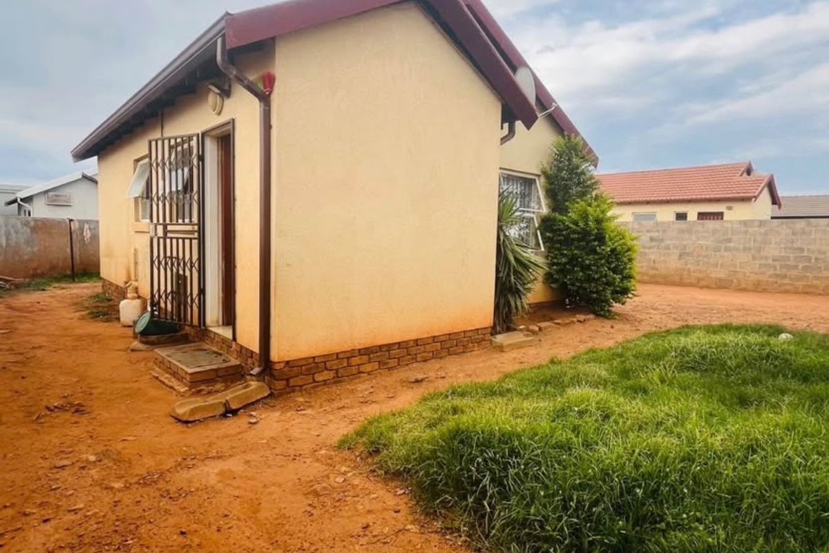 Rear view of a detached house featuring a pitched roof, metal security gate, and an unpaved yard area with a concrete boundary wall.