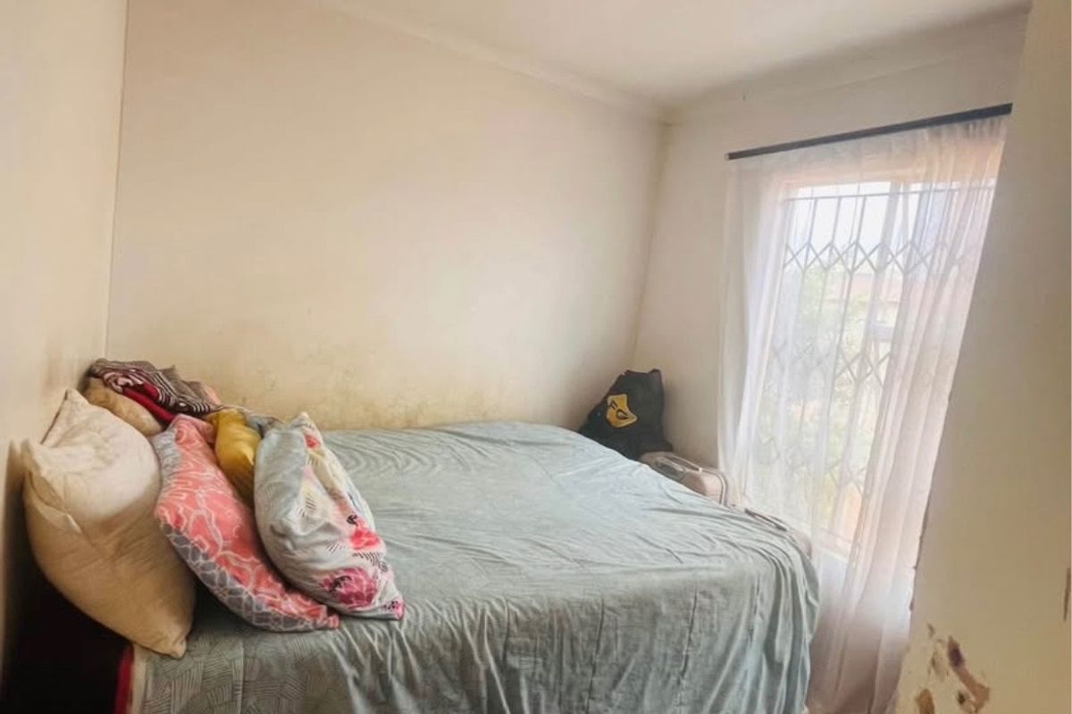 A bedroom featuring a window with a security gate and curtain rod, showing visible wall damage and staining requiring maintenance and cosmetic repair.