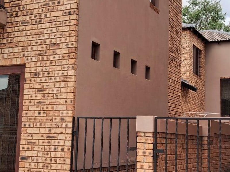 Exterior view of a townhouse featuring a combination of face brick and plastered walls, with a paved driveway and secure wrought iron gate entrance.