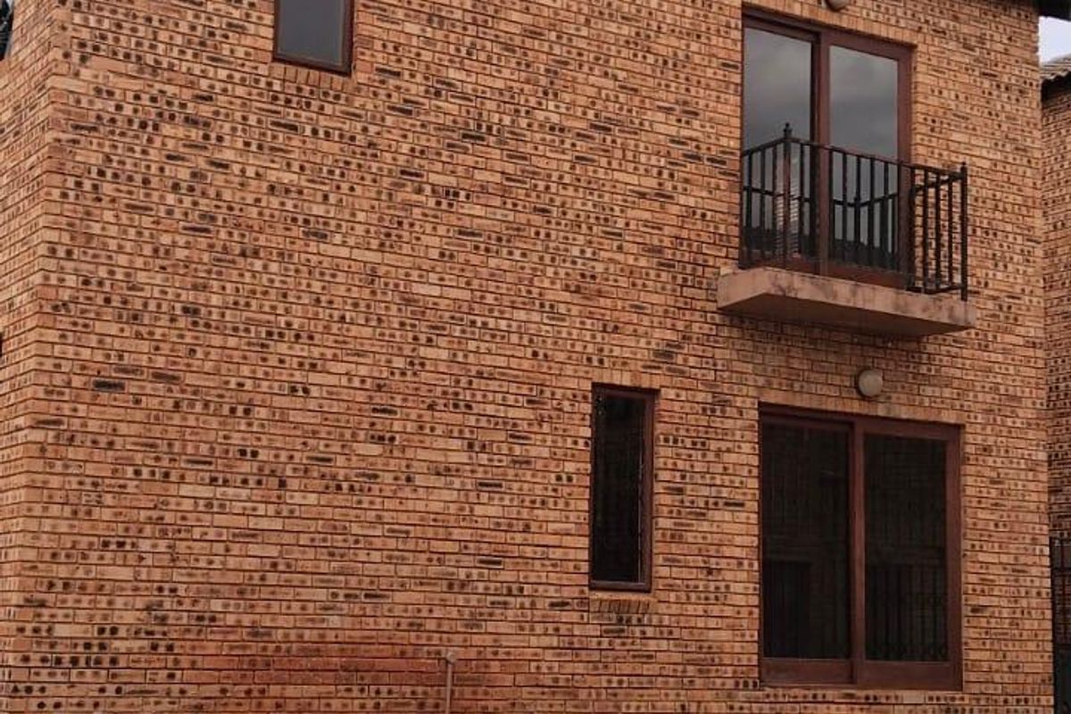 A two-story townhouse exterior featuring face brick construction, a small balcony with wrought iron railing, and a paved driveway area.