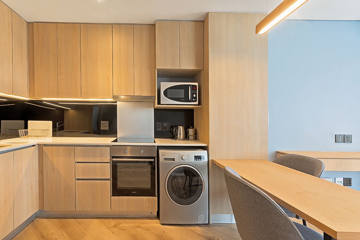 Modern kitchen featuring light timber cabinetry, integrated appliances including an oven, hob, and washing machine, complemented by a sleek mirrored splashback and breakfast bar.