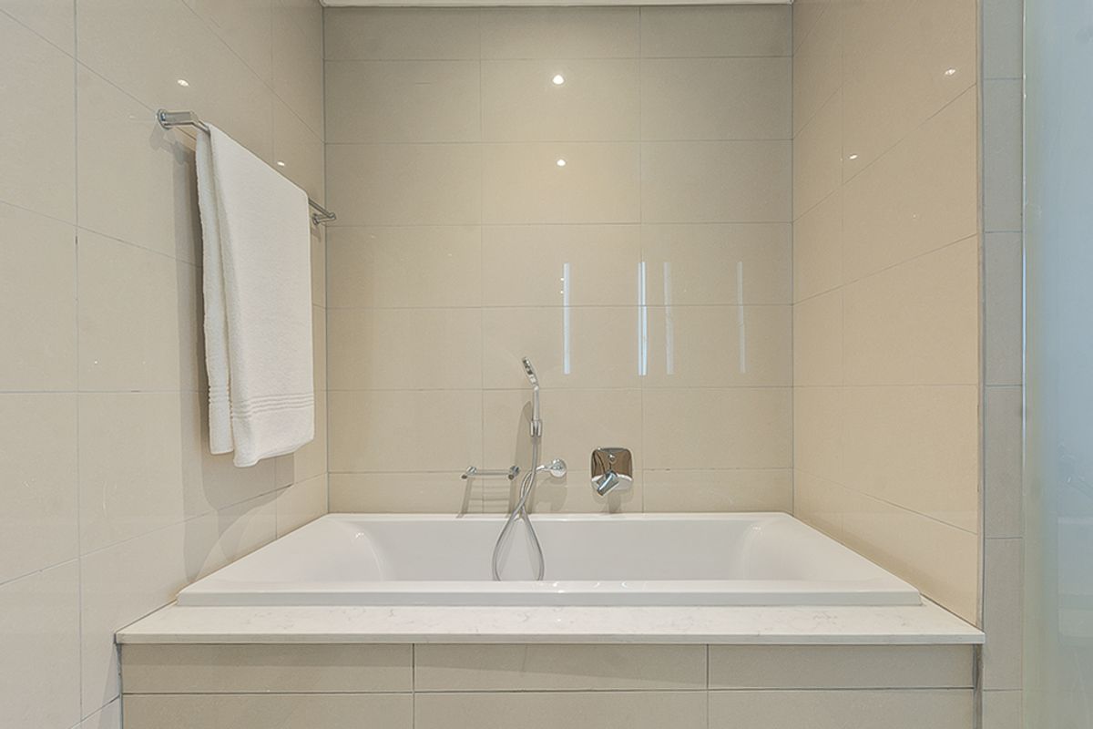 A clean, modern bathroom featuring a built-in bathtub with chrome fixtures, neutral wall tiling, and a stone-topped surround.
