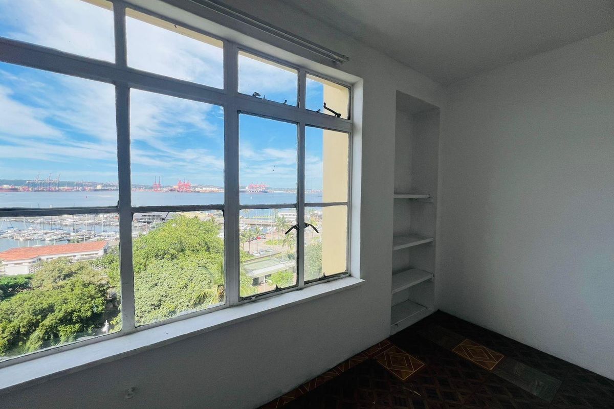 A bright bedroom featuring original parquet flooring, built-in shelving, and large windows offering expansive harbor and city views.