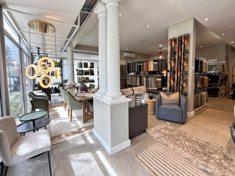 A well-lit, open-plan commercial showroom space featuring structural columns, polished concrete floors, and large glass windows, currently utilized for interior design displays.