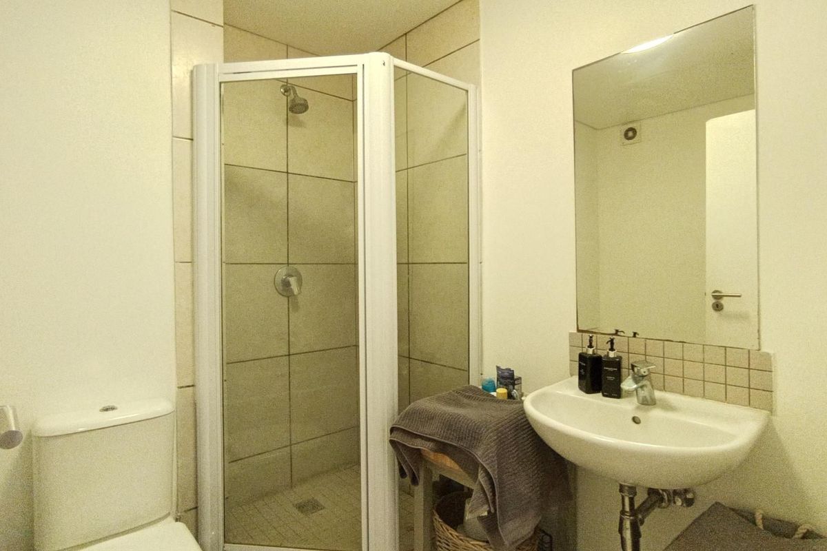 A functional bathroom featuring a corner shower enclosure, pedestal washbasin, and wall-mounted mirror with neutral wall tiling.