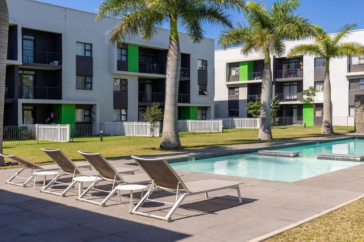 A well-maintained communal swimming pool area featuring a paved deck, landscaped garden, and modern apartment building exteriors in the background.
