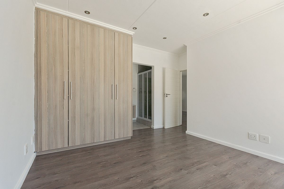 A bright, empty bedroom featuring modern wood-look laminate flooring, built-in cupboards, and recessed downlighting, with an adjacent en-suite bathroom visible.