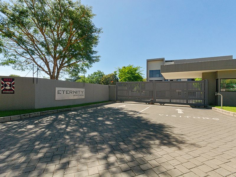 The secure entrance to the Eternity Rivonia apartment complex, featuring a paved visitor driveway, automated security gate, and perimeter boundary wall with electric fencing.