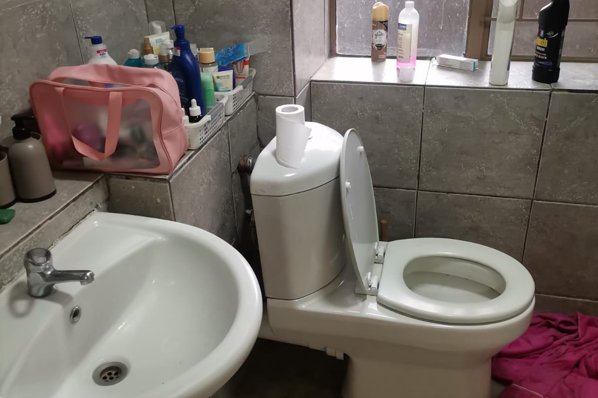 A functional bathroom featuring a pedestal basin, a standard toilet, and grey wall and floor tiling with a window providing natural light.