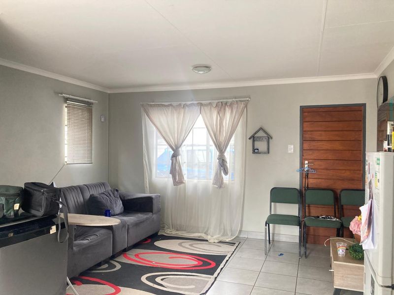 A functional living room featuring tiled flooring, a timber entrance door, and natural light through window treatments.