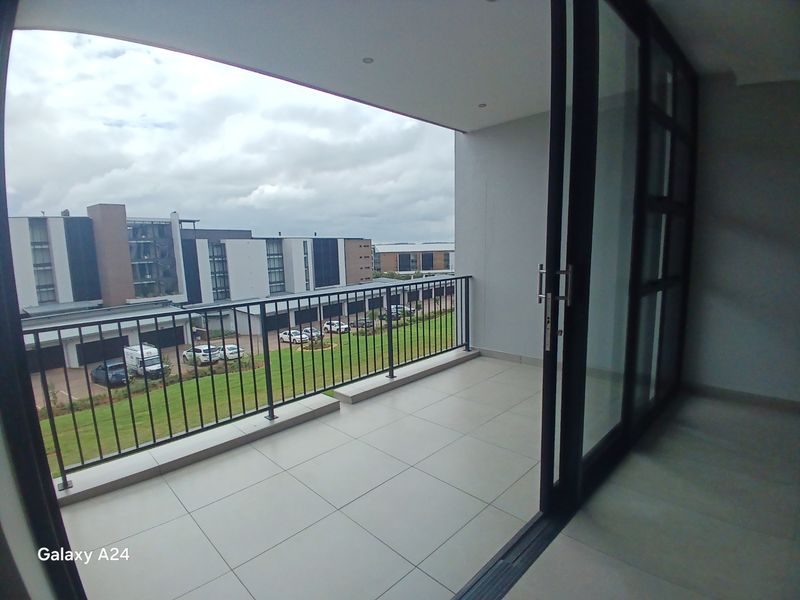 A modern, covered balcony featuring tiled flooring, a sleek metal balustrade, and large sliding glass doors, offering an open view of the surrounding estate.
