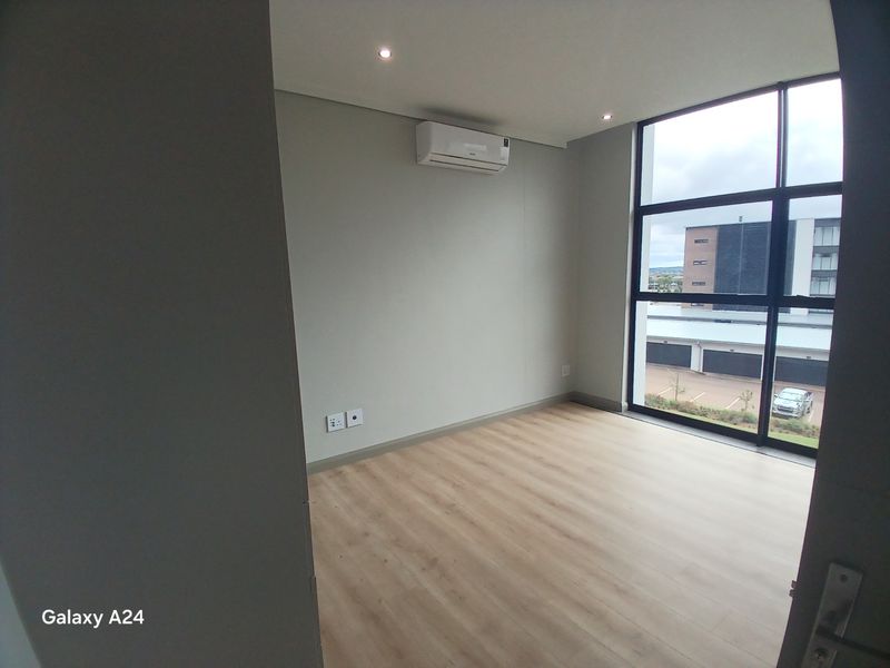 A bright, empty bedroom featuring modern laminate flooring, a wall-mounted air conditioning unit, and large floor-to-ceiling windows with natural light.