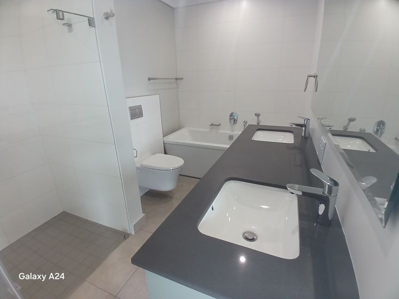 Modern bathroom featuring a double vanity with dark stone countertop, wall-hung toilet, built-in bathtub, and a spacious walk-in shower with neutral tiling.