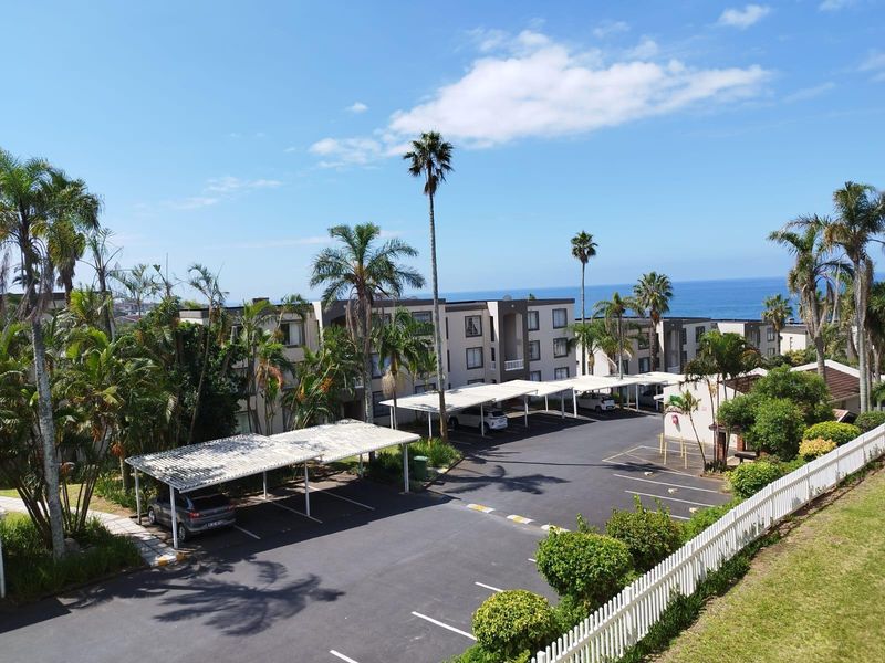 Aerial view of the apartment complex parking area featuring covered carports, landscaped grounds, and a white picket fence with ocean views in the background.
