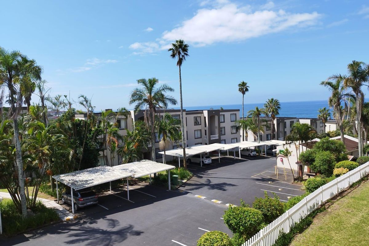 Aerial view of the apartment complex parking area featuring covered carports, landscaped grounds, and a white picket fence with ocean views in the background.
