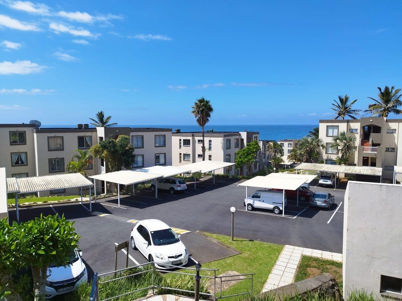 Elevated view of the apartment complex exterior, featuring paved parking areas with covered carports, landscaped grounds, and ocean views in the background.