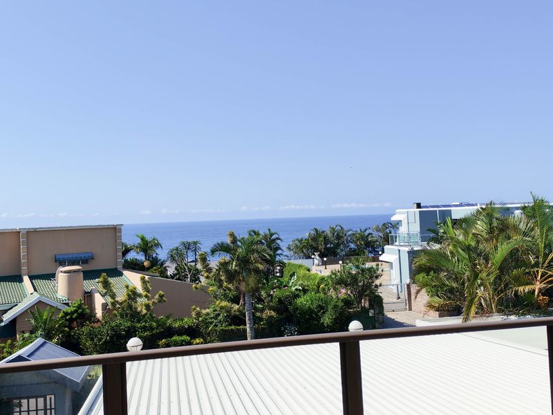 A scenic elevated view overlooking lush coastal vegetation, neighbouring properties, and the ocean horizon from a balcony.