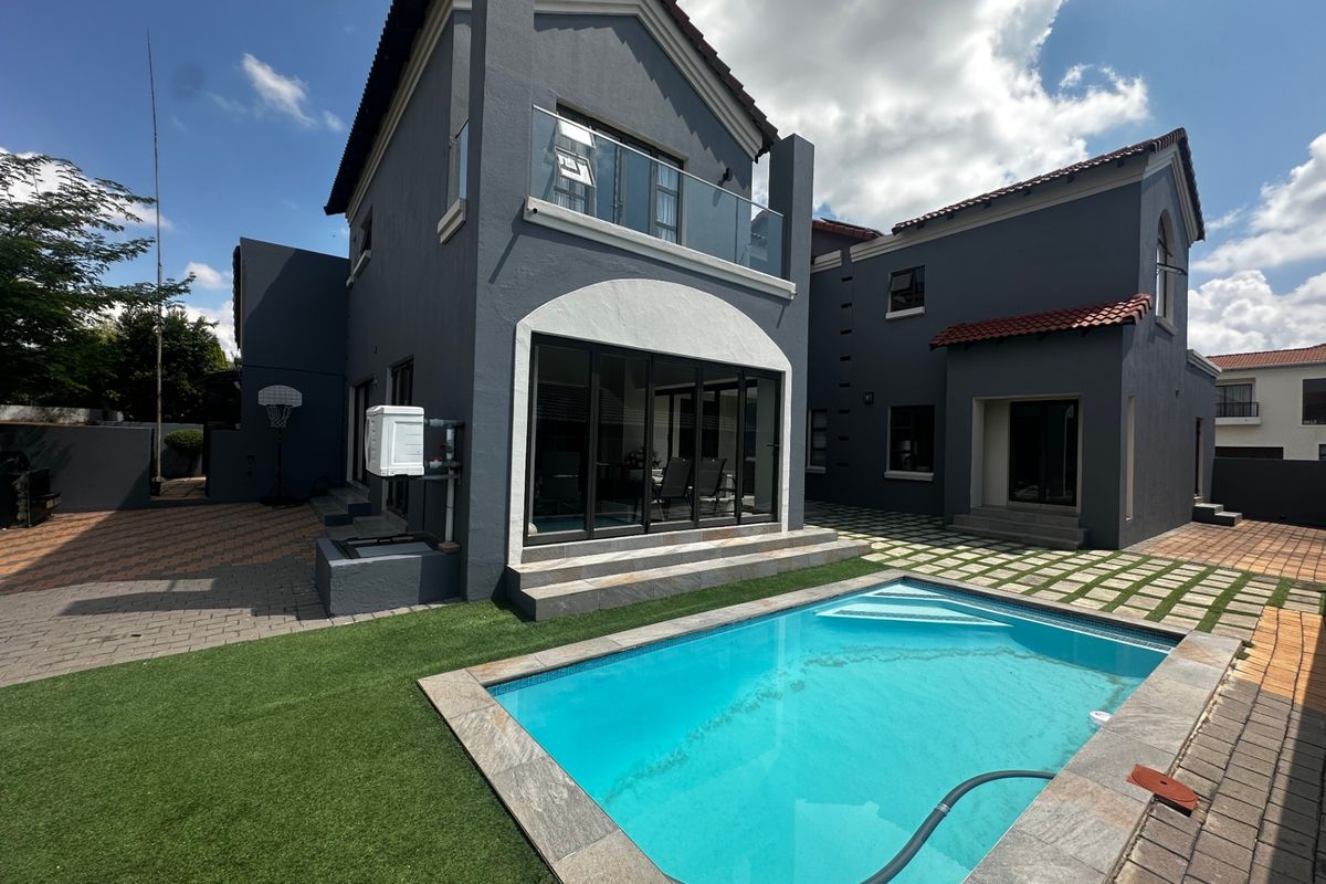 Rear view of a modern two-story house featuring a swimming pool, paved patio area, artificial lawn, and a glass-railed balcony.