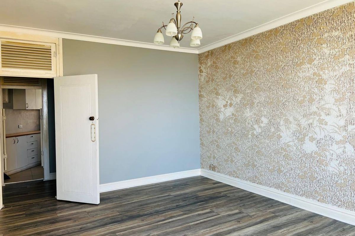 An empty living room featuring laminate flooring, decorative wallpaper, and a central chandelier, with an adjacent view into the kitchen area.
