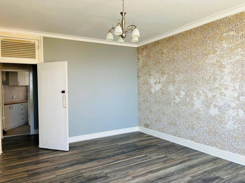 An empty living room featuring laminate flooring, decorative wallpaper, and a central chandelier, with an adjacent view into the kitchen area.