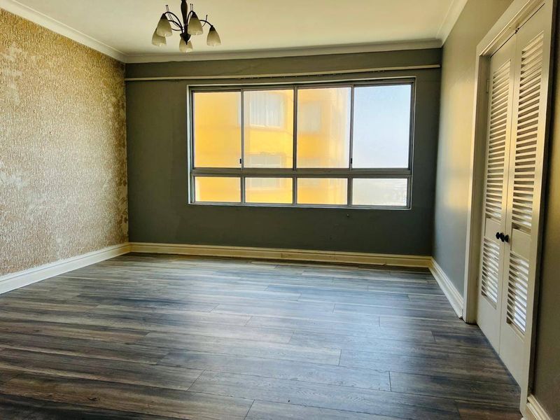 An empty bedroom featuring laminate flooring, louvered closet doors, and a large window. The walls require cosmetic repair and refinishing.