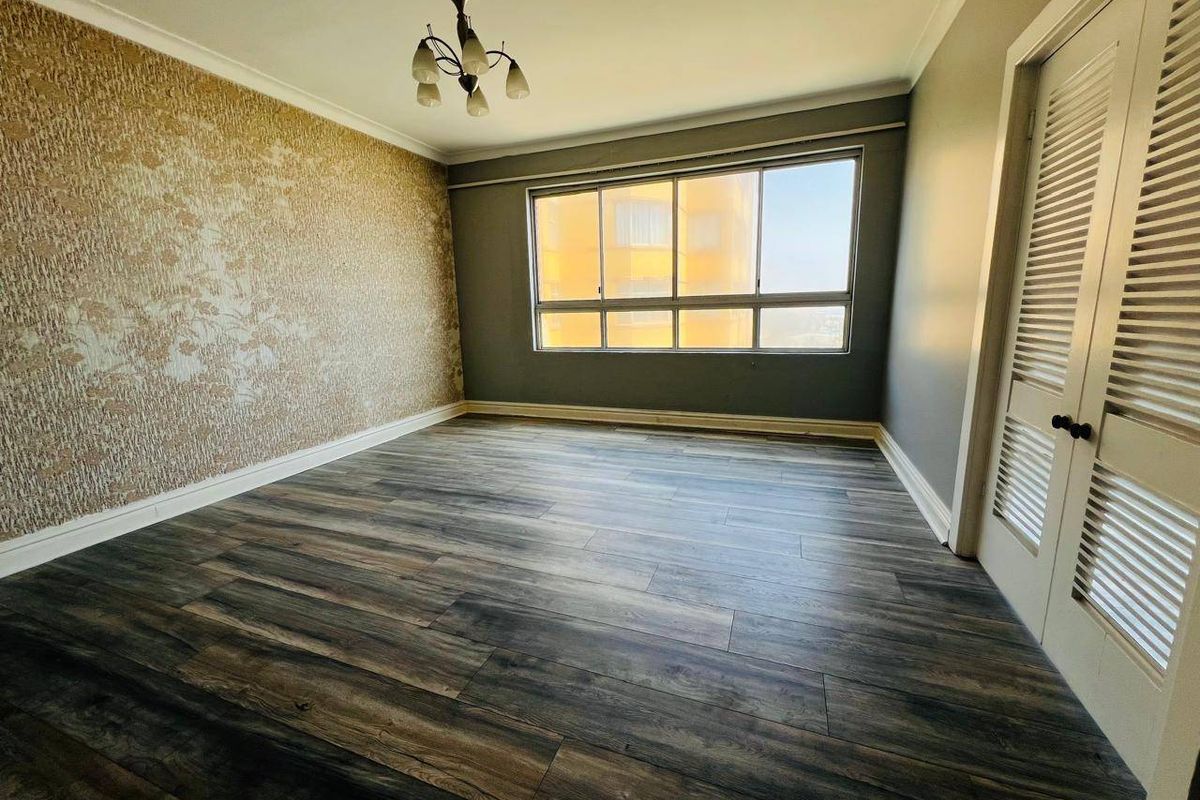 A bright, empty bedroom featuring modern laminate flooring, a feature wallpaper wall, and built-in cupboards with louvered doors.