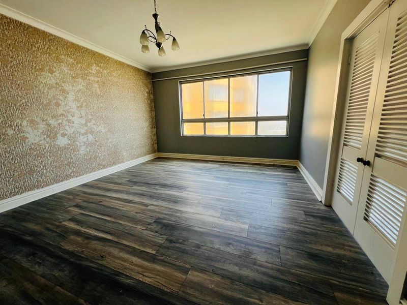 A bright, empty bedroom featuring modern laminate flooring, a feature wallpaper wall, and built-in cupboards with louvered doors.