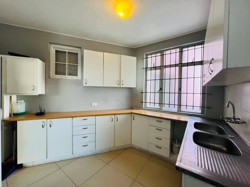 A functional kitchen featuring white built-in cabinetry, laminate countertops, a double stainless steel sink, and tiled flooring with security bars on the window.