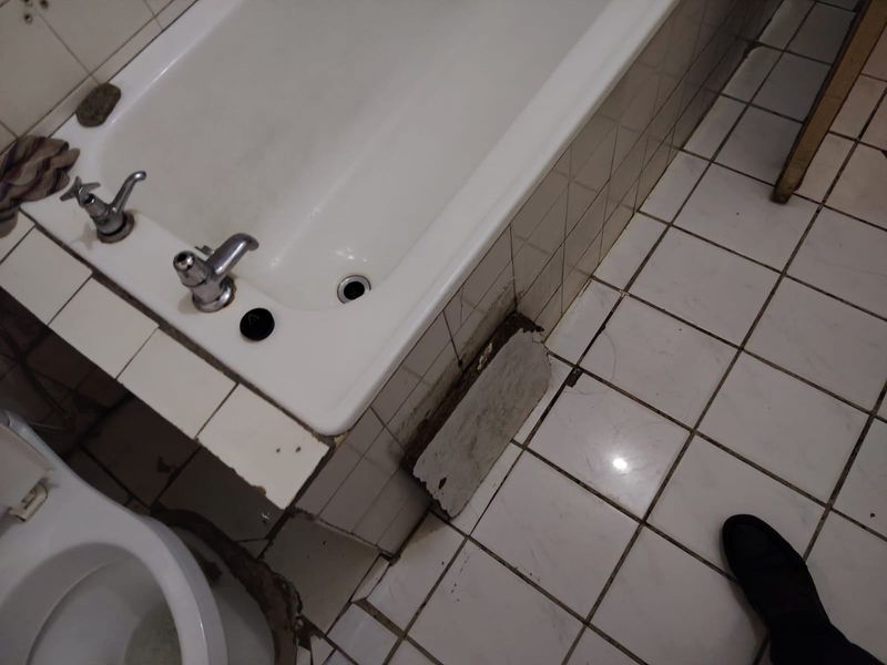 A bathroom featuring a bathtub and tiled flooring, currently showing significant wear and damage to the bath surround and tiling.