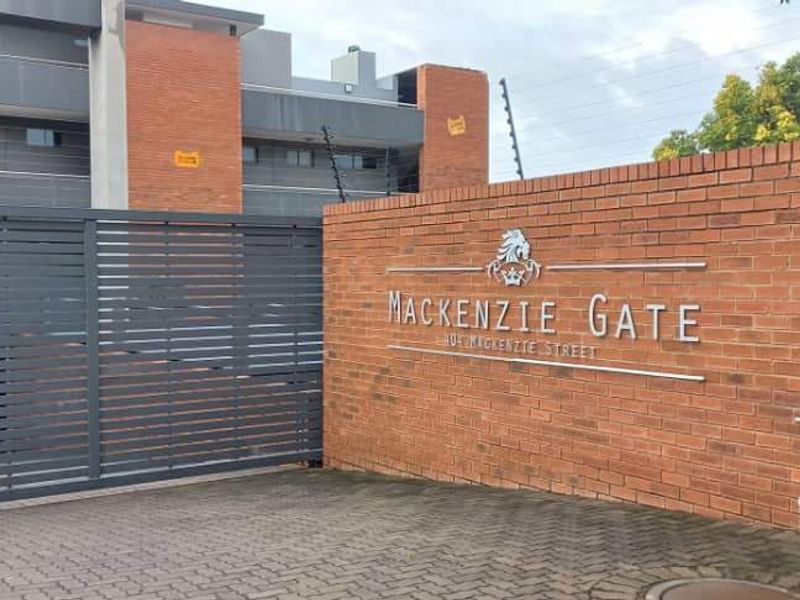 Front entrance view of Mackenzie Gate apartment complex featuring a brick boundary wall, secure sliding gate, and paved driveway access.