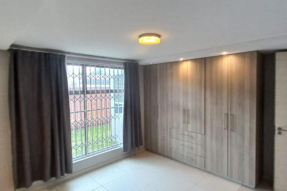 A bright, empty bedroom featuring modern built-in cupboards, tiled flooring, and a window fitted with a security gate.