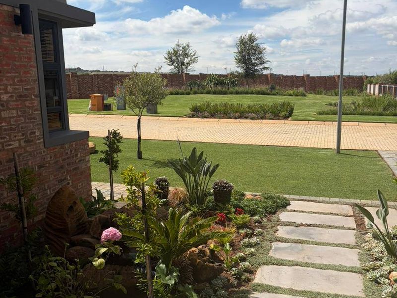 A well-maintained garden featuring a paved walkway, landscaped beds, and a brick exterior facade, overlooking a spacious lawn and boundary wall.
