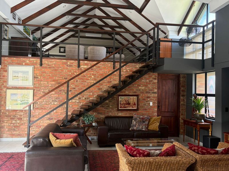Spacious living area featuring a double-volume ceiling with exposed timber trusses, a striking brick feature wall, and a mezzanine level accessed by a steel staircase.