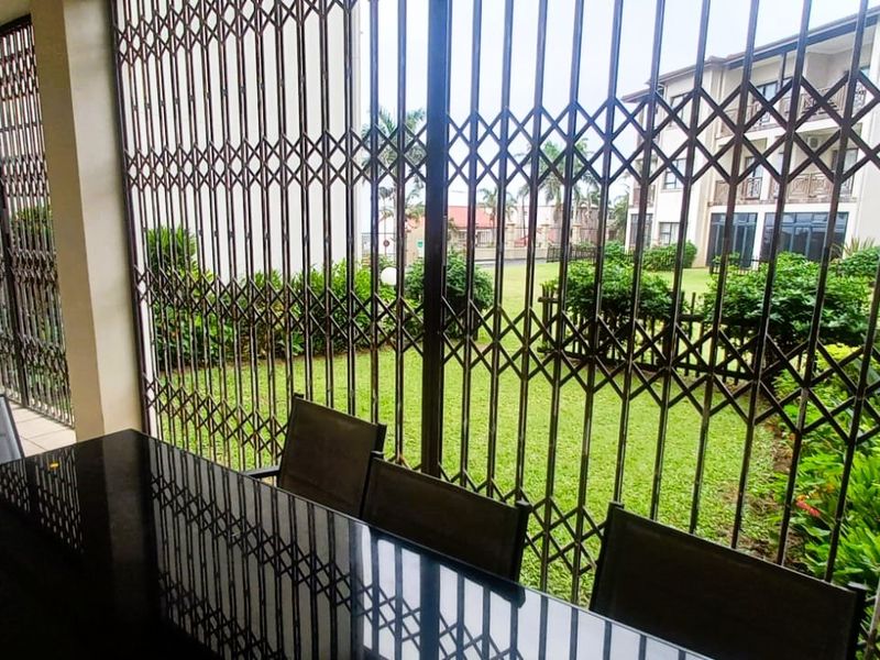 A covered patio area featuring security gates, tiled flooring, and an outdoor air conditioning unit, overlooking a communal garden space.