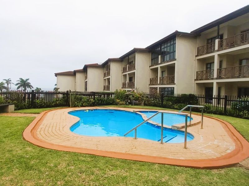 Communal swimming pool area set within a landscaped garden, featuring paved surrounds and views of the apartment complex exterior.