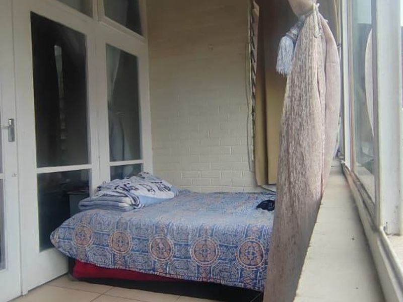 A narrow enclosed balcony or sunroom featuring tiled flooring, large windows, and a painted brick wall, currently utilized as a sleeping area.