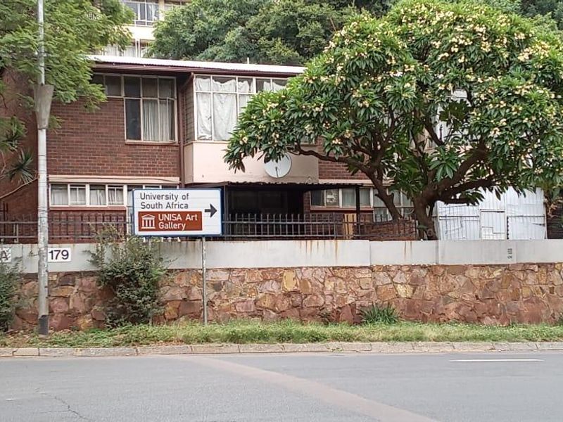 Street-facing view of a brick apartment building featuring a stone-clad boundary wall, metal security fencing, and mature landscaping.