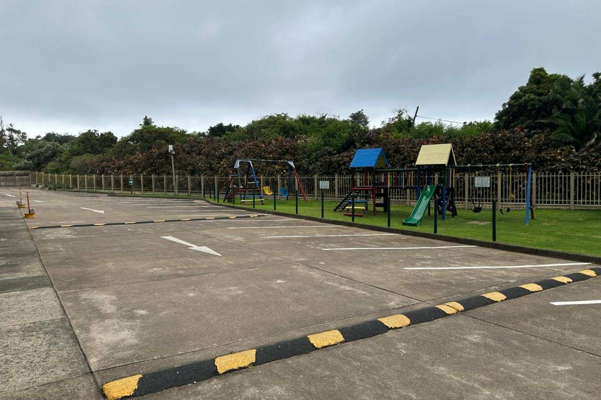 A paved parking area featuring marked bays, speed bumps, and an adjacent communal children's playground enclosed by metal fencing.