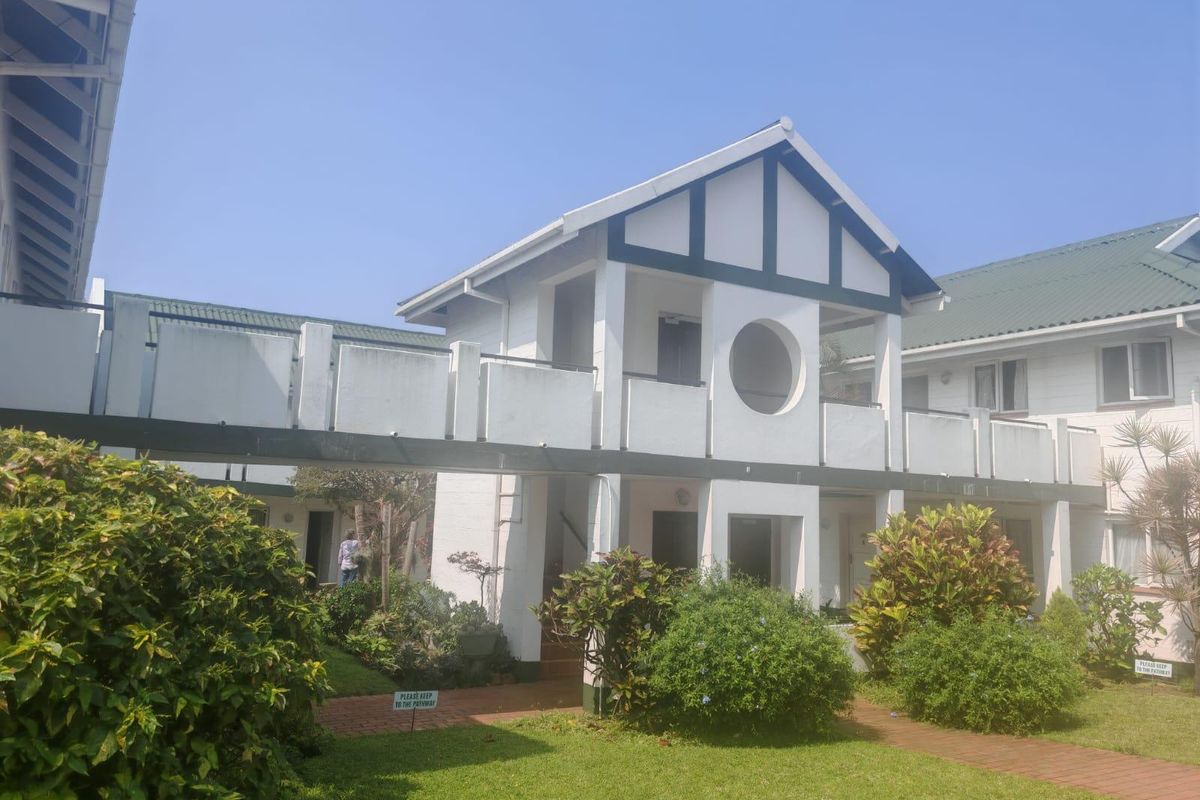 Exterior view of an apartment complex featuring a white facade with timber detailing, balconies, and a well-maintained garden area with brick pathways.