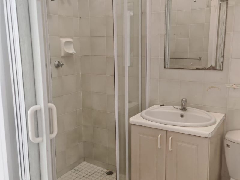 A functional bathroom featuring a shower enclosure, vanity cabinet with basin, and wall-mounted mirror, showing signs of wear and dated finishes.