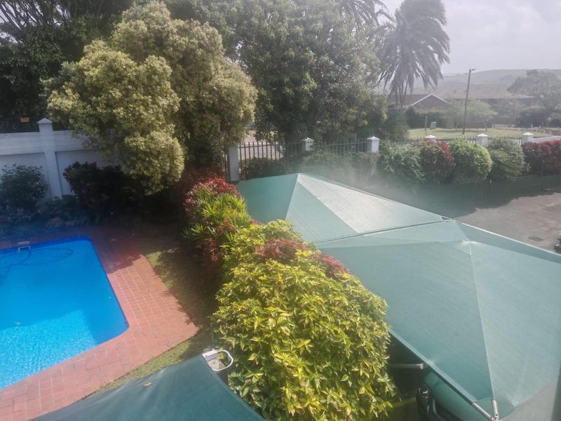 An elevated view of a residential swimming pool area featuring brick coping, mature landscaping, and a large green shade sail structure.