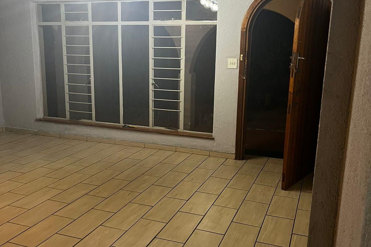 An empty living room featuring tiled flooring, a distinctive arched wooden door, and a large window with security bars.