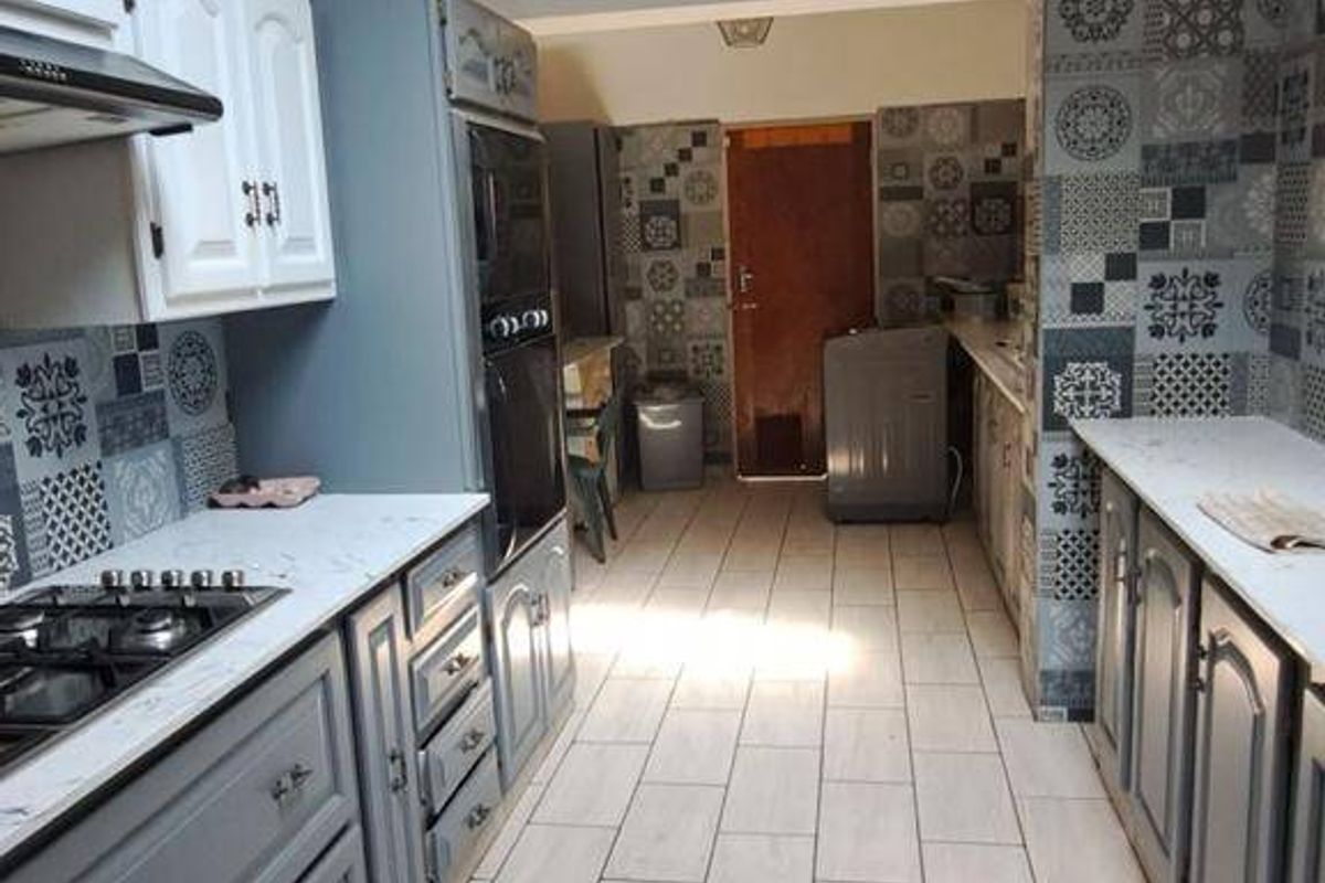 A galley-style kitchen featuring grey cabinetry, a gas hob, built-in oven, decorative tiled backsplash, and exposed ceiling beams.
