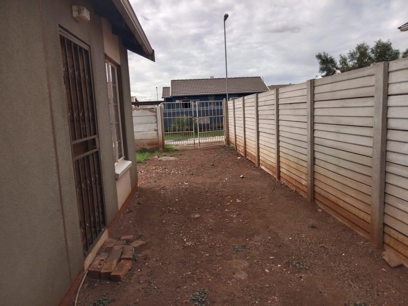 A side or rear yard area featuring a precast concrete boundary wall, metal security gate, and unlandscaped ground surface.