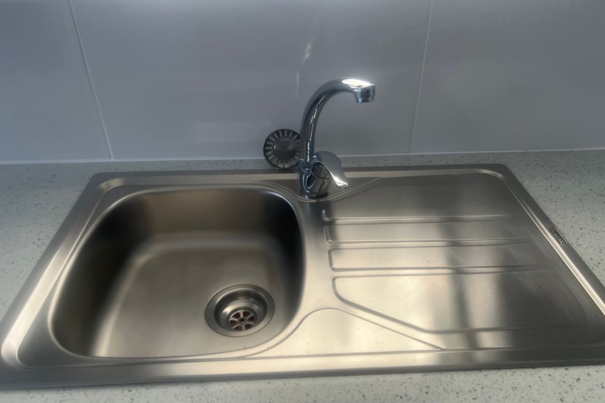 A clean stainless steel kitchen sink with a modern chrome mixer tap, set into a speckled stone-look countertop with white tiled splashback.