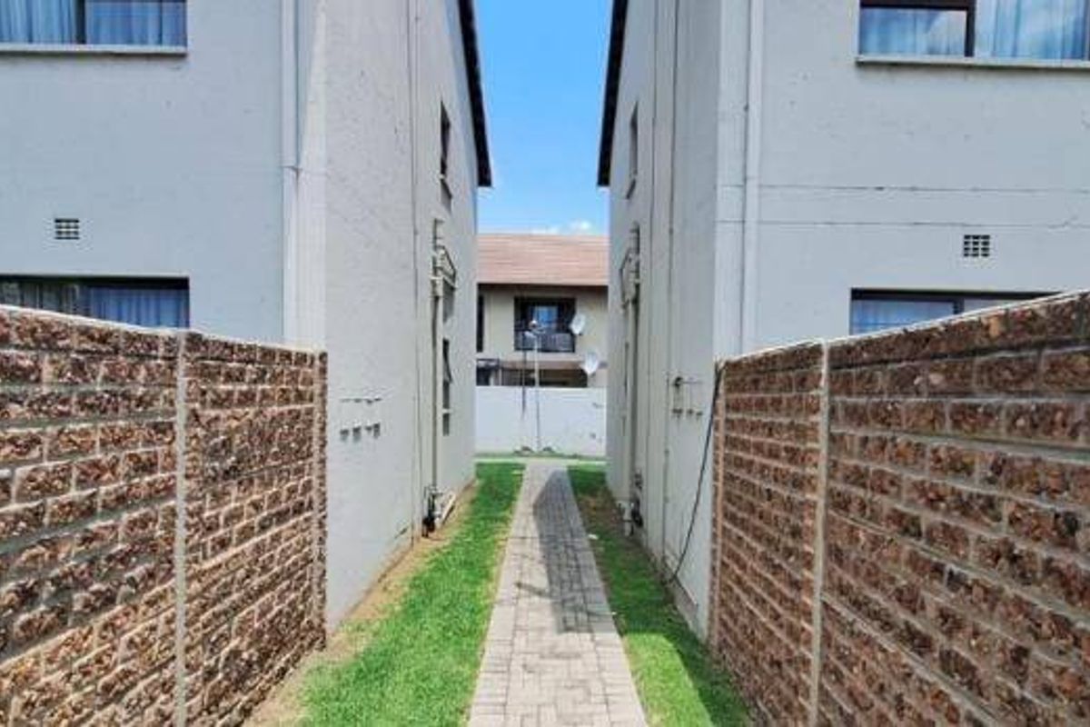 A view of the exterior walkway between apartment blocks, featuring brick boundary walls, a paved path, and grass landscaping.