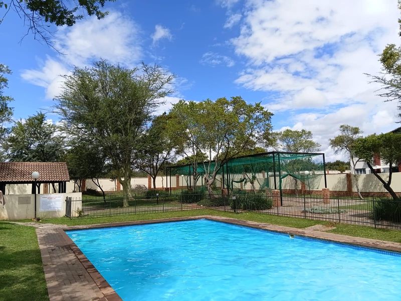 Communal swimming pool area featuring a paved surround, covered gazebo, and landscaped garden within a secure fenced perimeter.